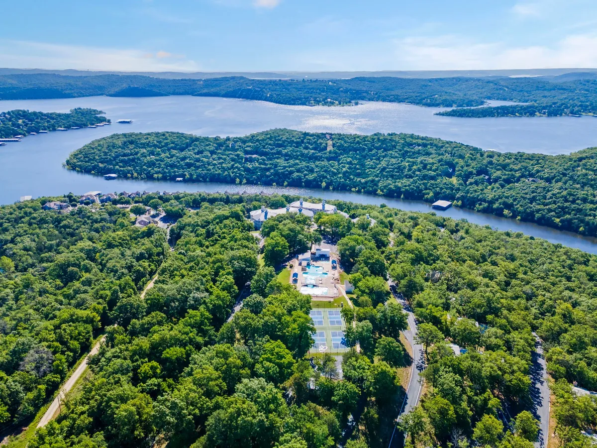 An expansive lake view with lush greenery, a swimming pool, and a tennis court are all visible. The setting captures the tranquil yet sociable vibe of the resort, with the evening sun hinting at memorable gatherings by the water or around a fire pit.