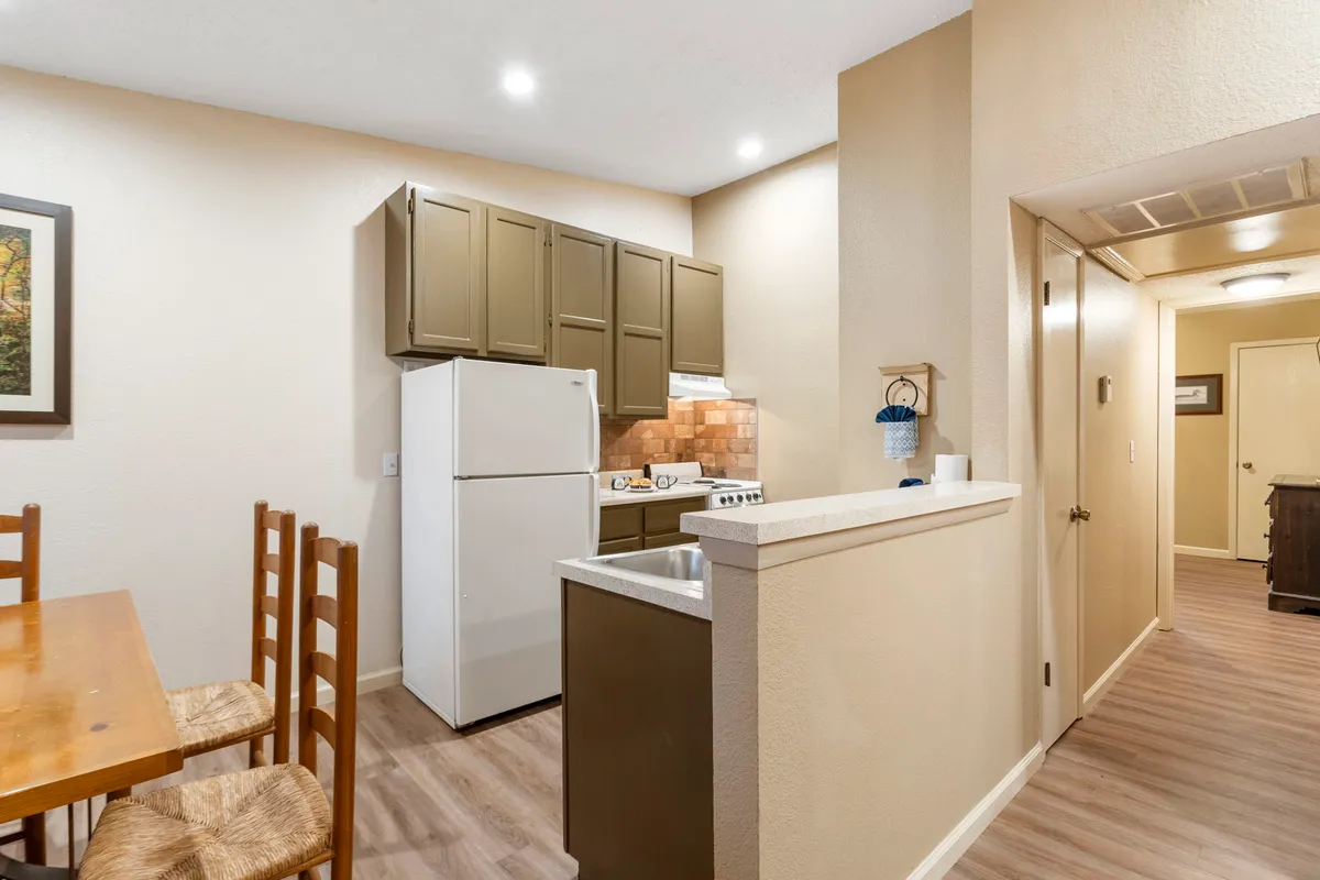 A spacious, homey interior showing both the kitchen cabinets and dining table, with a refrigerator and stove visible in the background. The scene suggests meals being prepared and shared, with the table ready for gatherings.