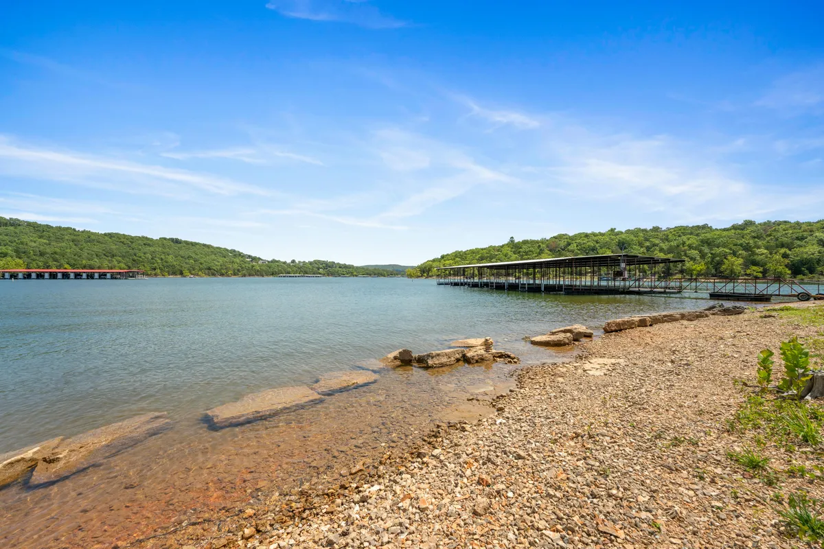 A serene view across calm lake waters, highlighting a sturdy dock set against a rocky shoreline and surrounded by lush greenery. The scene invites fishing, boating, and relaxed nature appreciation, with the landscape emphasizing the easy access to the water from the property.