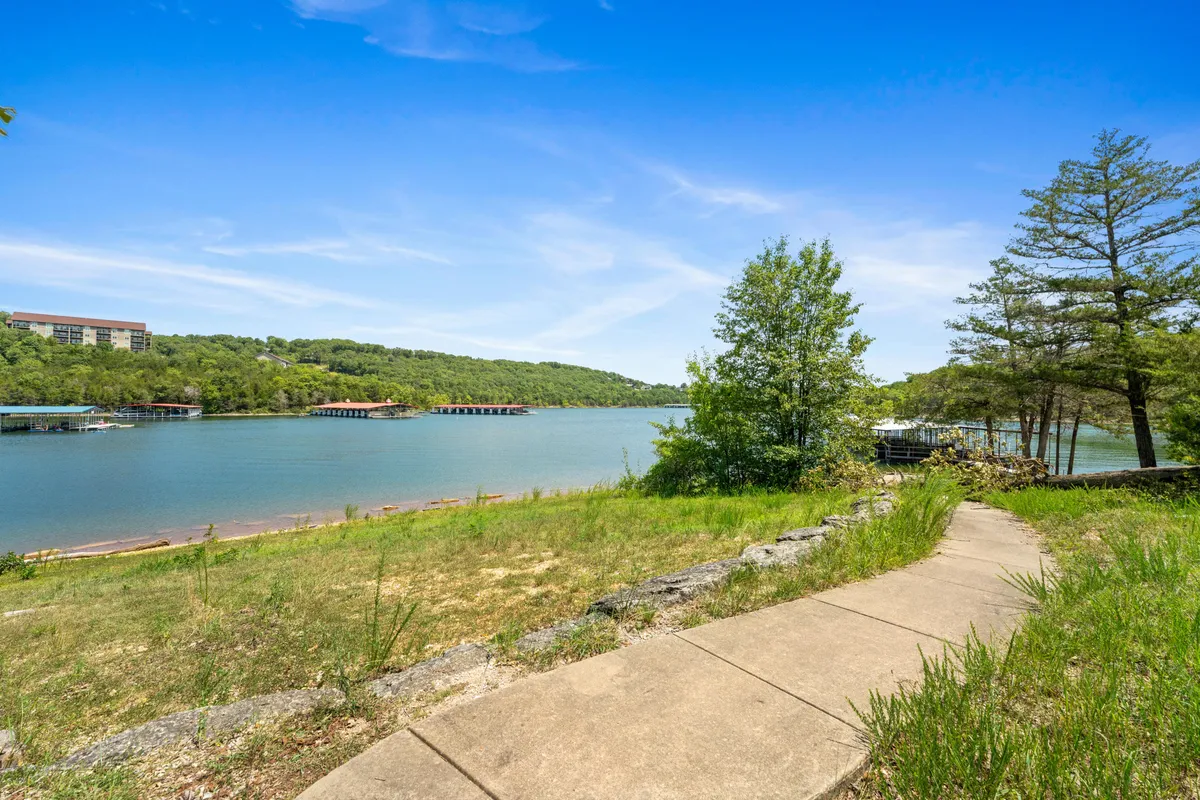 A tranquil waterfront scene with a winding pathway bordered by lush greenery, gentle hills in the distance, and calm water stretching out from the edge. The path invites leisurely walks and is surrounded by nature, perfect for strolls with pets.
