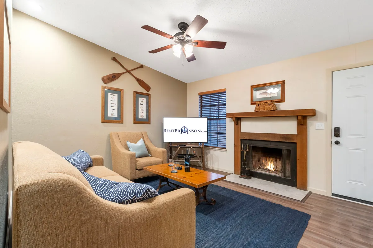 A warm, inviting living room featuring a plush sofa set arranged in front of a stone fireplace, a coffee table for games or snacks, and a wall-mounted television. Decorative oars and rustic accents enhance the cozy, lakeside ambiance. Soft lighting and the arrangement suggest a perfect gathering spot for both lively game nights and quiet evenings.