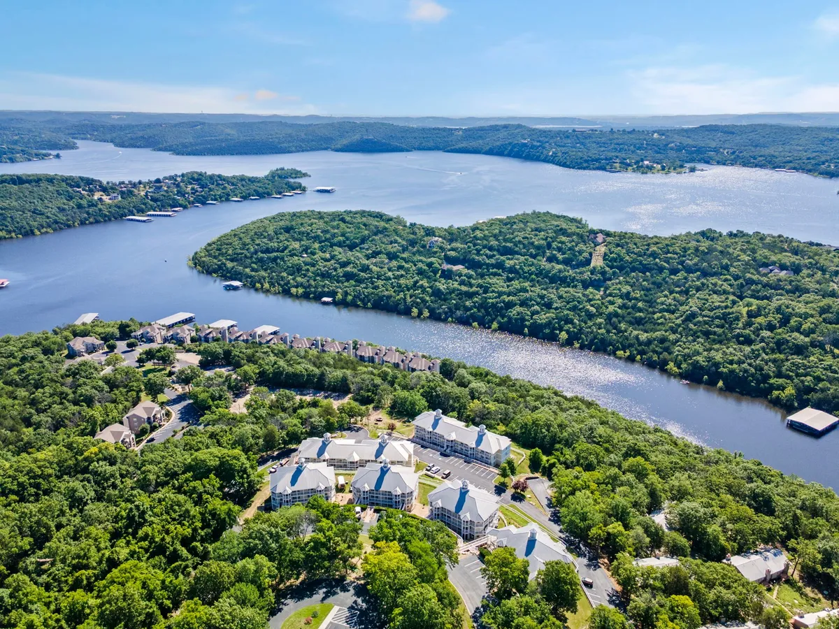 A scenic view of a wide river bordered by an island, wooded areas, and some residential buildings in the distance. The area is bathed in the warm light of sunset, with gentle ripples on the water and hints of people boating or fishing. The landscape feels peaceful and inviting, emphasizing natural beauty and outdoor activities.