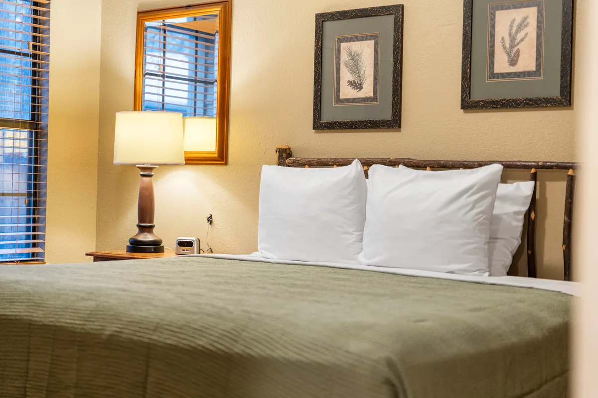 A tranquil bedroom featuring a bed with crisp white pillows, a softly lit bedside lamp, and framed artwork on the wall. Natural light streams in from a nearby window, giving the space a serene and restful atmosphere.