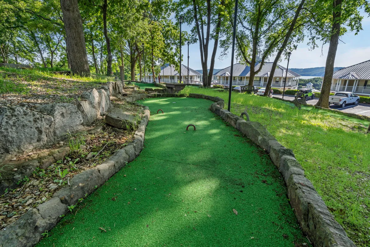 This image showcases a vibrant mini-golf putting green surrounded by lush trees and stone pathways, with nearby resort buildings visible in the background. The scene exudes a joyful family-friendly atmosphere, inviting guests to enjoy outdoor games and picnics together.