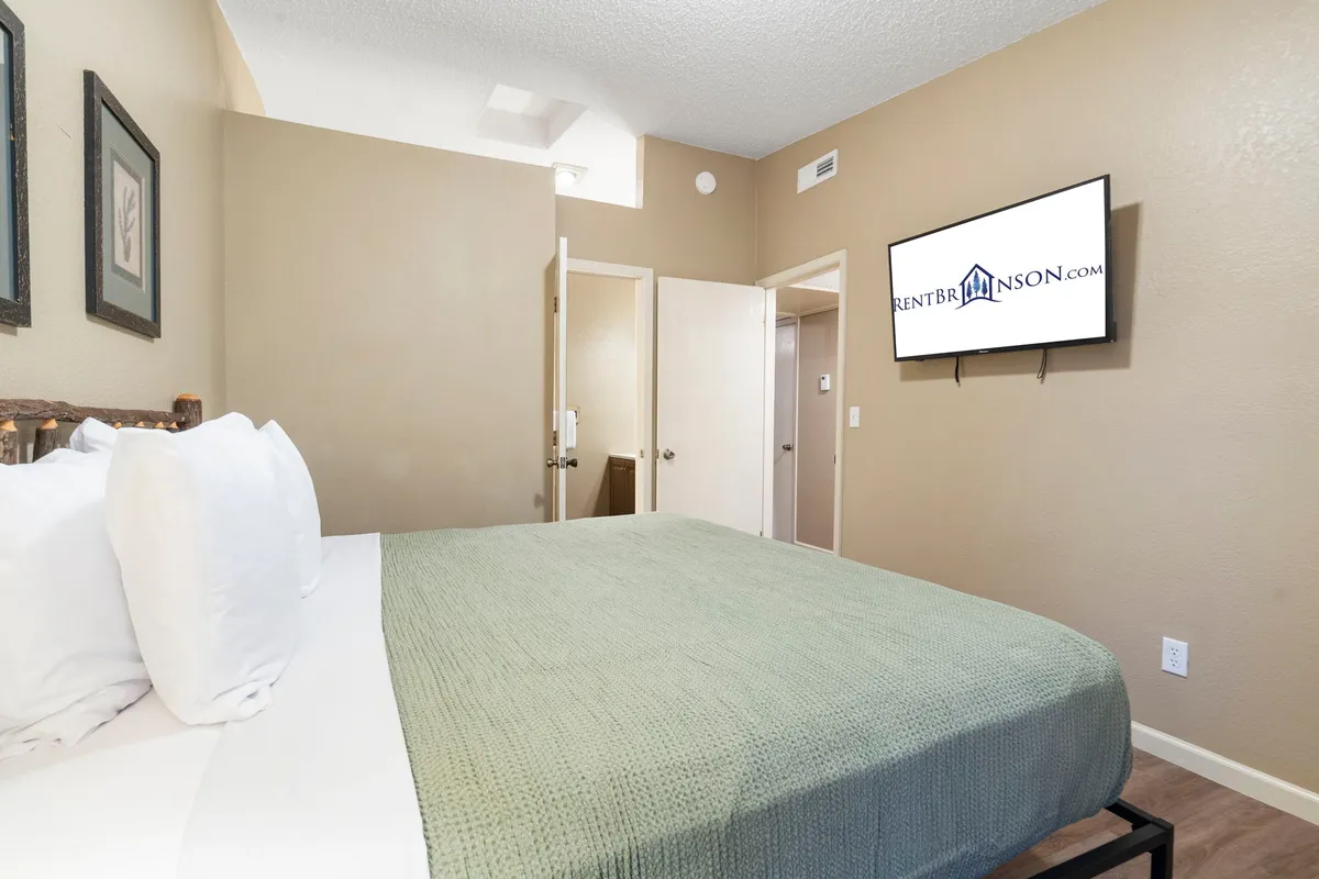 A calm, contemporary bedroom with a green textured blanket on the bed, a wall-mounted TV, and well-chosen artwork. The room’s soft tones and spacious feel create a restful environment for guests.