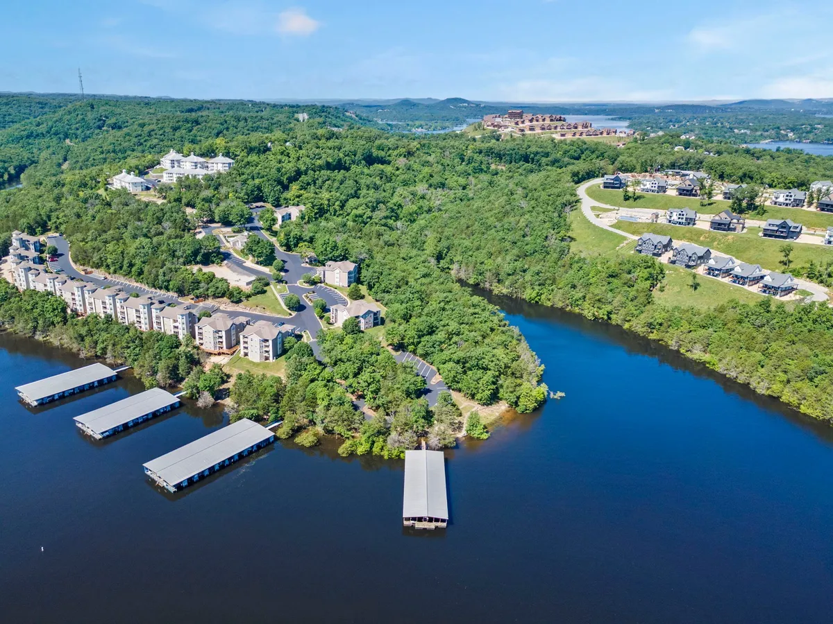 Lakefront view featuring wooded hills, residential buildings, and boat docks along the water's edge. The scene is peaceful, with ample open space and access to both the lake and natural surroundings.